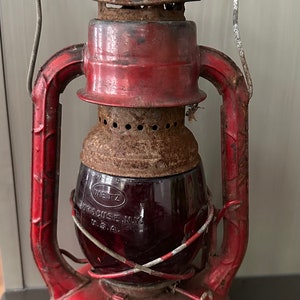 May include: Vintage red Dietz lantern with a clear glass globe. The lantern has a metal handle and is marked "Syracuse N.Y. U.S.A." The lantern shows signs of age and wear, with rust and paint loss. A classic piece of Americana.