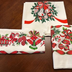 May include: Three folded white Christmas-themed textiles with red and green designs. The designs include bells, bows, ornaments, candy canes, and pinecones. The textiles are arranged on a dark brown wooden surface.