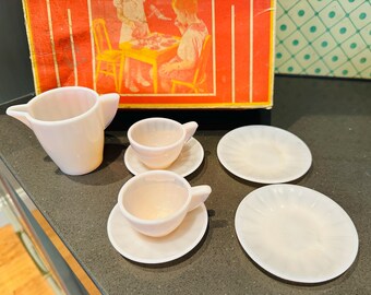 May include: A vintage pink toy tea set, including a creamer, two teacups with saucers, and two plates. The set is made of a translucent material and is displayed in front of a box with a retro illustration.