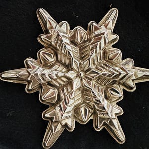May include: A detailed, silver-colored snowflake ornament. The ornament has a symmetrical, multi-pointed design with intricate patterns and textures. The snowflake is set against a dark background, highlighting its reflective surface and detailed craftsmanship.