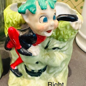 May include: A vintage ceramic planter featuring a whimsical elf figure with a green cap, blue eyes, and a red bow tie. The planter has a light green, textured exterior with a smiling face design. The elf is dressed in black and red.