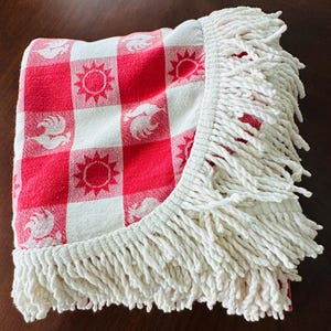 May include: A folded red and white checkered picnic blanket with a white fringe border. The blanket features repeating sun and rooster designs within the checkered pattern. The fringe is made of thick, twisted white yarn.