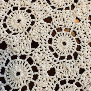 May include: An off-white crochet doily featuring a repeating circular motif pattern. The intricate needlework consists of interconnected round centers surrounded by openwork stitching, creating a delicate, lace-like texture throughout the fabric.