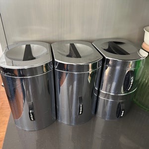 May include: Three shiny, chrome-colored kitchen canisters with black labels that read "FLOUR", "SUGAR", and "COFFEE". The canisters have a rectangular shape with rounded corners and a black handle on the lid. A vintage mixer and glass jar are in the background.
