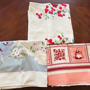 May include: Three folded rectangular fabric cloths. One features a red cherry pattern on a cream background. Another displays blue and pink floral motifs on a light blue and cream base. The third shows a red grid pattern with floral and figure designs.