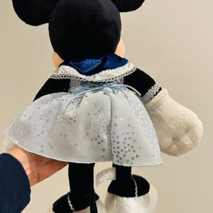 K&ouml;nnte beinhalten: Eine Minnie Mouse Pl&uuml;schpuppe, von hinten gesehen, tr&auml;gt ein hellblaues Kleid mit silbernen Glitzerakzenten. Die Puppe hat schwarze Ohren, eine blaue Schleife und wei&szlig;e Handschuhe und Schuhe. Das Kleid hat einen blauen Samtkragen.