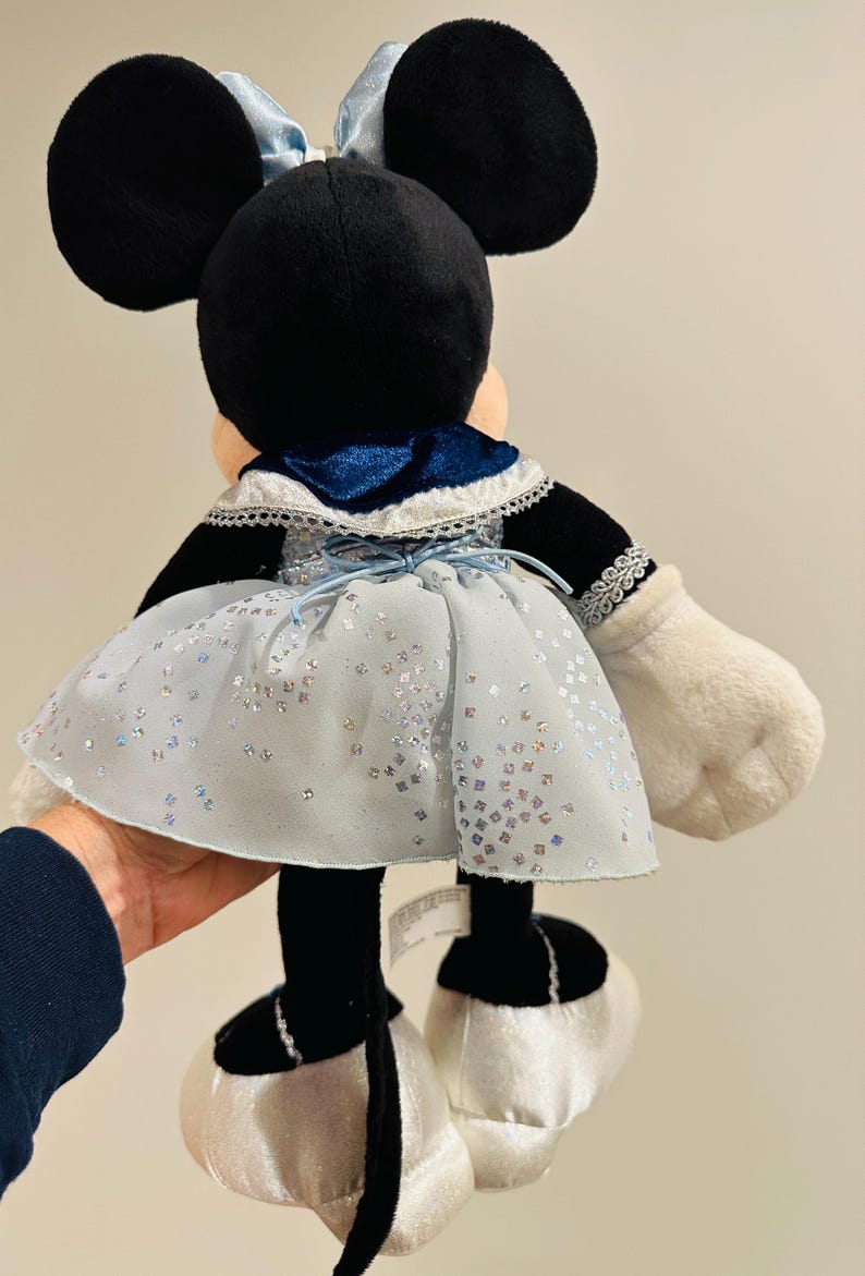 K&ouml;nnte beinhalten: Eine Minnie Mouse Pl&uuml;schpuppe, von hinten gesehen, tr&auml;gt ein hellblaues Kleid mit silbernen Glitzerakzenten. Die Puppe hat schwarze Ohren, eine blaue Schleife und wei&szlig;e Handschuhe und Schuhe. Das Kleid hat einen blauen Samtkragen.