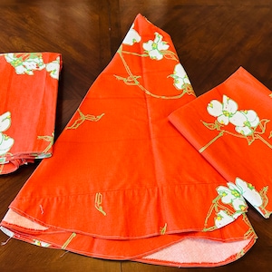 May include: A set of orange fabric items, including a folded rectangle, a triangular shape, and a square, all featuring a white floral pattern with green accents. The items are arranged on a dark wooden surface.