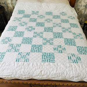 May include: White quilt with a teal geometric pattern. The quilt features a repeating block design, with each block composed of teal rectangles arranged in a grid. The quilt is placed on a wooden bed frame.