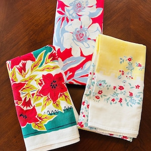 May include: Three folded rectangular kitchen towels. One features a vibrant pink background with large white and light blue floral patterns. Another displays red flowers with green leaves on a white base. The third has a yellow border with small red and blue floral accents.