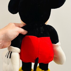 May include: A plush Mickey Mouse toy, viewed from the back. The toy features a black head and ears, a black body, red shorts, white gloves, and yellow shoes. The tail is black. The toy is soft and cuddly.