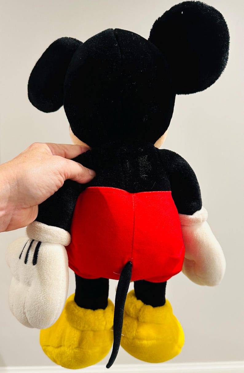 May include: A plush Mickey Mouse toy, viewed from the back. The toy features a black head and ears, a black body, red shorts, white gloves, and yellow shoes. The tail is black. The toy is soft and cuddly.