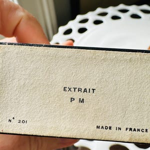 K&ouml;nnte beinhalten: Nahaufnahme einer Vintage-Box mit dem Text "EXTRAIT P M" und "MADE IN FRANCE". Die Box ist beige mit schwarzem Rand und der Nummer "201" aufgedruckt. Die Box wird in einer Hand gehalten.