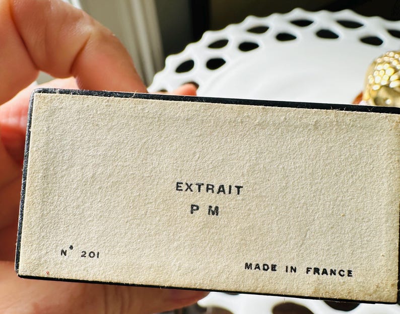 K&ouml;nnte beinhalten: Nahaufnahme einer Vintage-Box mit dem Text "EXTRAIT P M" und "MADE IN FRANCE". Die Box ist beige mit schwarzem Rand und der Nummer "201" aufgedruckt. Die Box wird in einer Hand gehalten.