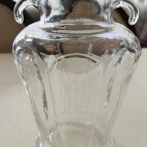 May include: Clear glass vase with a black lid. The vase has two curved handles and a wide, rounded body. The glass is transparent, allowing a view of the interior. The vase is likely used for decorative purposes or holding flowers.