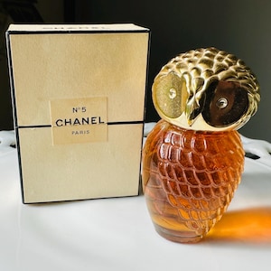 K&ouml;nnte beinhalten: Ein Vintage-Parf&uuml;mflakon von Chanel No. 5 in Form einer Eule, mit einem goldfarbenen Kopf und einem strukturierten, bernsteinfarbenen K&ouml;rper. Daneben eine beige Schachtel mit dem Chanel-Logo und Text.