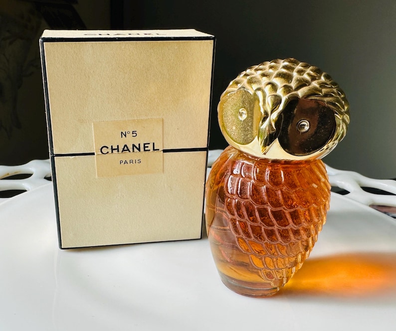 K&ouml;nnte beinhalten: Ein Vintage-Parf&uuml;mflakon von Chanel No. 5 in Form einer Eule, mit einem goldfarbenen Kopf und einem strukturierten, bernsteinfarbenen K&ouml;rper. Daneben eine beige Schachtel mit dem Chanel-Logo und Text.