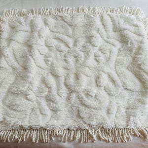 May include: Cream-colored rectangular bath mat with a textured floral pattern. The mat has a fringe border. The soft, plush material is visible in this close-up shot. This decorative mat is suitable for a bathroom.