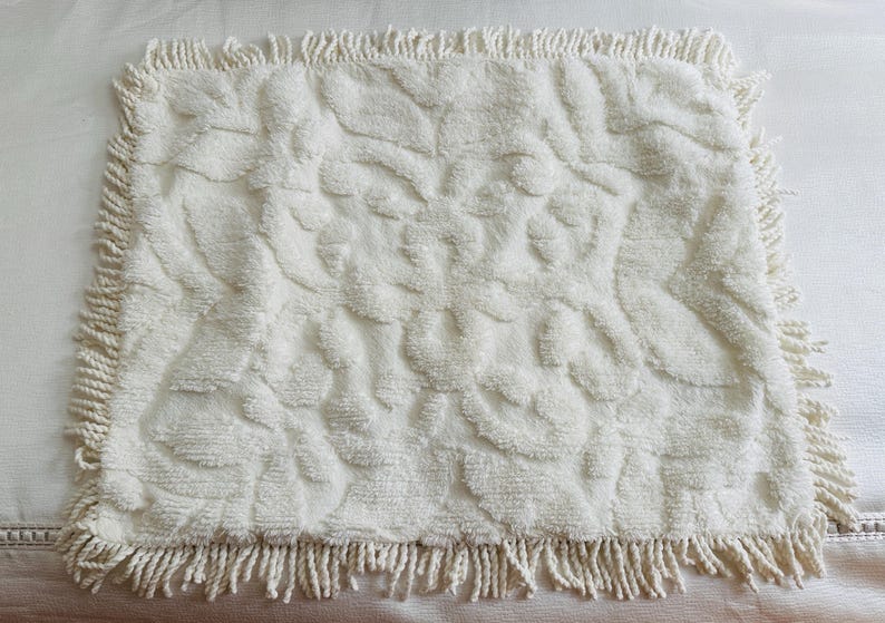 May include: Cream-colored rectangular bath mat with a textured floral pattern. The mat has a fringe border. The soft, plush material is visible in this close-up shot. This decorative mat is suitable for a bathroom.
