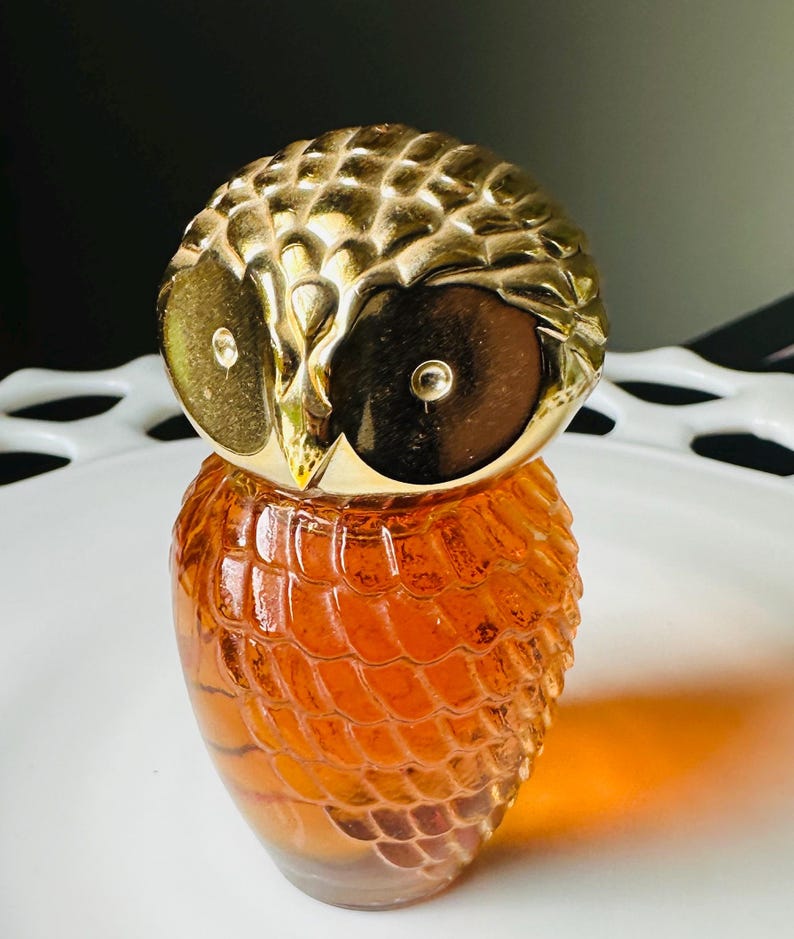 K&ouml;nnte beinhalten: Eine bernsteinfarbene Glasparf&uuml;mflasche in Form einer Eule. Die Flasche hat ein strukturiertes, schuppenartiges Muster. Der Eulenkopf ist aus goldfarbenem Metall mit detaillierten Merkmalen, einschlie&szlig;lich gro&szlig;er, dunkler Augen.