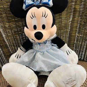 K&ouml;nnte beinhalten: Ein Pl&uuml;sch Minnie Mouse Puppe mit schwarzen Ohren, einem rosa Gesicht und einer blauen Schleife. Sie tr&auml;gt ein hellblaues Kleid mit silbernen Akzenten und wei&szlig;en Handschuhen. Die Puppe sitzt auf einem Korbstuhl.