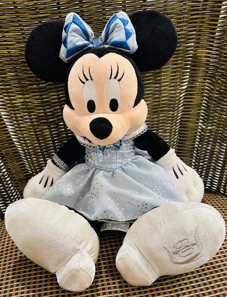 K&ouml;nnte beinhalten: Ein Pl&uuml;sch Minnie Mouse Puppe mit schwarzen Ohren, einem rosa Gesicht und einer blauen Schleife. Sie tr&auml;gt ein hellblaues Kleid mit silbernen Akzenten und wei&szlig;en Handschuhen. Die Puppe sitzt auf einem Korbstuhl.