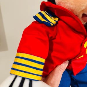 May include: A plush toy wearing a red jacket with blue and yellow striped accents on the sleeves and collar. The toy has blue pants and white gloves with black stripes. The toy is held by a person.