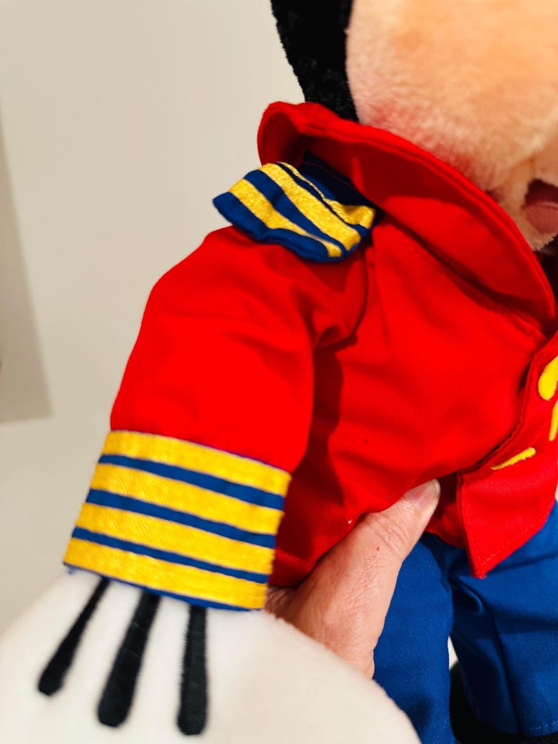 May include: A plush toy wearing a red jacket with blue and yellow striped accents on the sleeves and collar. The toy has blue pants and white gloves with black stripes. The toy is held by a person.