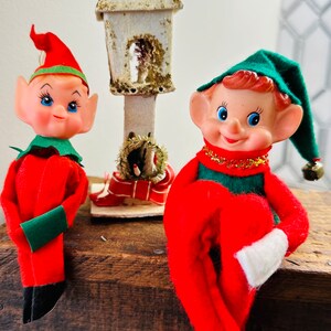 May include: Two vintage Christmas elf figurines with red suits, green accents, and elf hats. One elf has a red hat, the other a green hat. A small, white, glitter-covered birdhouse is between them. The elves are on a wooden surface.