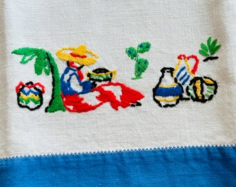 May include: Embroidered white linen towel with a blue border. The embroidery depicts a person in a sombrero, seated under a palm tree, with colorful picnic items. The design features red, yellow, blue, green, and black thread.