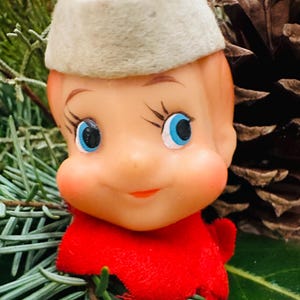 May include: A vintage Christmas elf figurine with a rosy complexion, blue eyes, and a red outfit. The elf wears a felt hat with blue accents. The figurine is surrounded by evergreen branches and a pine cone.