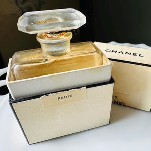 K&ouml;nnte beinhalten: Vintage Chanel No. 5 Parf&uuml;mflakon mit einem transparenten, geometrischen Verschluss. Die Flasche befindet sich in ihrer Originalverpackung, mit dem Chanel-Logo und dem Wort "Paris" sichtbar. Die Box ist cremefarben mit schwarzem Rand.