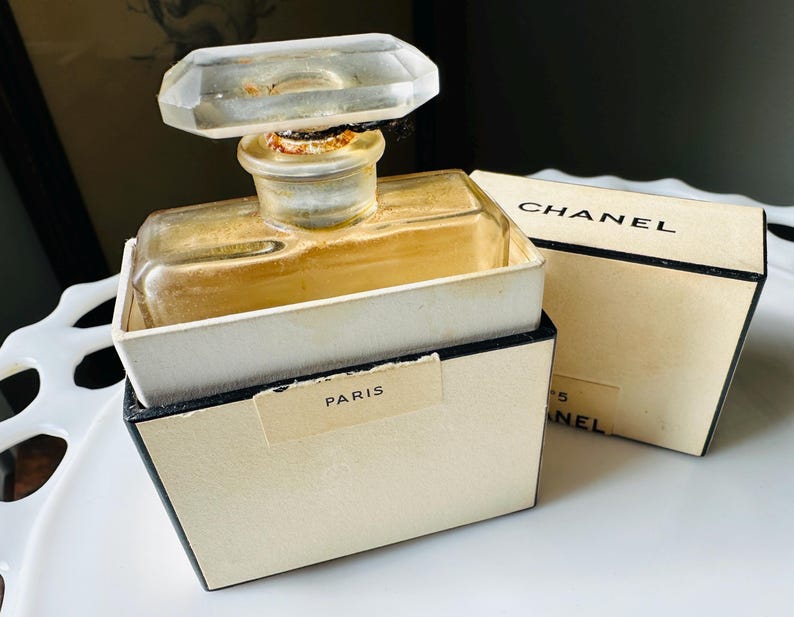 K&ouml;nnte beinhalten: Vintage Chanel No. 5 Parf&uuml;mflakon mit einem transparenten, geometrischen Verschluss. Die Flasche befindet sich in ihrer Originalverpackung, mit dem Chanel-Logo und dem Wort "Paris" sichtbar. Die Box ist cremefarben mit schwarzem Rand.