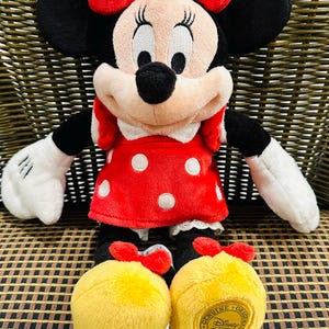Puede incluir: Mu&ntilde;eca de peluche de Minnie Mouse, con un vestido rojo con lunares blancos, un lazo a juego y zapatos amarillos. La mu&ntilde;eca tiene orejas negras, una nariz negra y guantes blancos. Los zapatos tienen un logotipo de Disney Store.