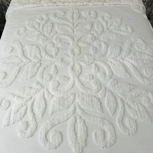 May include: White bedspread with a raised floral pattern. The design features a central medallion with stylized leaves and swirling details. The bedspread is made of a soft, textured material and is neatly arranged on a bed.