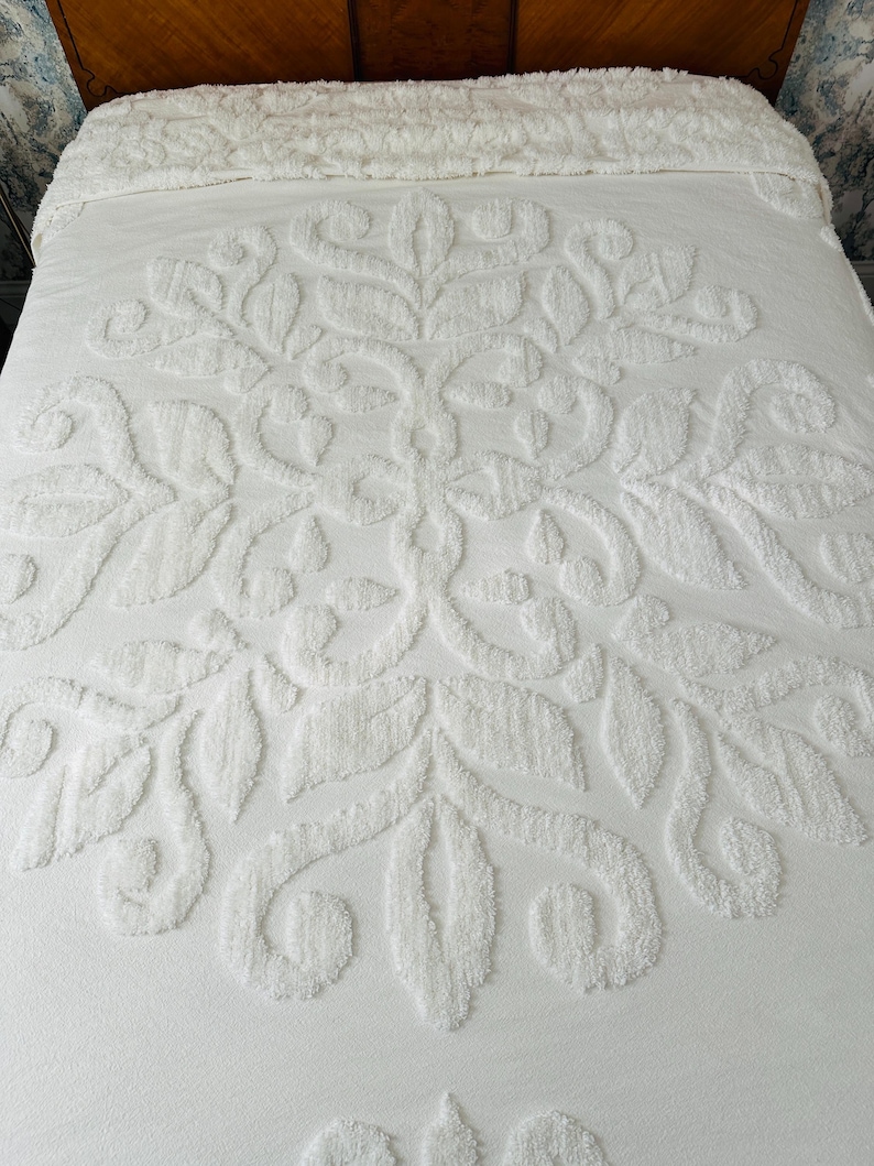 May include: White bedspread with a raised floral pattern. The design features a central medallion with stylized leaves and swirling details. The bedspread is made of a soft, textured material and is neatly arranged on a bed.