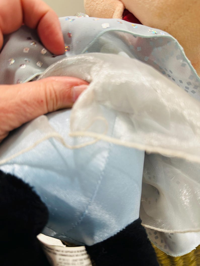 K&ouml;nnte beinhalten: Nahaufnahme einer Pl&uuml;schpuppe, die ein hellblaues Kleid mit silbernen Pailletten tr&auml;gt. Das Kleid hat einen wei&szlig;en, durchsichtigen &Uuml;berzug. Der schwarze Fu&szlig; der Puppe ist unten im Bild zu sehen.