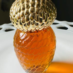K&ouml;nnte beinhalten: Eine dekorative Glasflasche mit einem goldfarbenen, strukturierten Deckel. Die Flasche hat einen bernsteinfarbenen Farbton und ein schuppenartiges Muster. Die Flasche steht auf einem wei&szlig;en, dekorativen Teller.