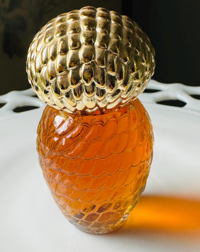 K&ouml;nnte beinhalten: Eine dekorative Glasflasche mit einem goldfarbenen, strukturierten Deckel. Die Flasche hat einen bernsteinfarbenen Farbton und ein schuppenartiges Muster. Die Flasche steht auf einem wei&szlig;en, dekorativen Teller.