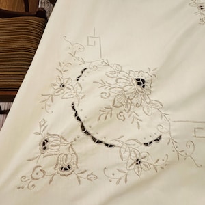 May include: Cream-colored tablecloth with intricate floral embroidery. The embroidery features detailed flowers and leaves in a complementary neutral color. The design is centered with a scalloped edge, adding a touch of elegance to the textile.
