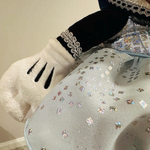 K&ouml;nnte beinhalten: Arm und Handschuh einer Pl&uuml;schfigur, verziert mit einem silbernen Armband. Die Figur tr&auml;gt ein hellblaues Kleid mit schimmernden, quadratischen Verzierungen. Der &Auml;rmel ist schwarz und der Handschuh wei&szlig; mit schwarzen Linien.