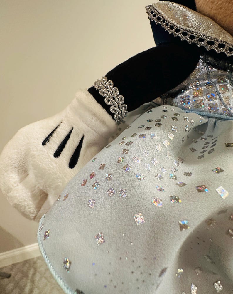 K&ouml;nnte beinhalten: Arm und Handschuh einer Pl&uuml;schfigur, verziert mit einem silbernen Armband. Die Figur tr&auml;gt ein hellblaues Kleid mit schimmernden, quadratischen Verzierungen. Der &Auml;rmel ist schwarz und der Handschuh wei&szlig; mit schwarzen Linien.