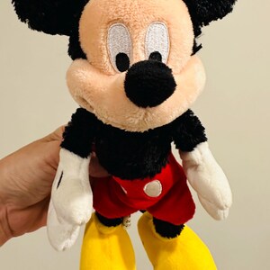 Puede incluir: Un peluche de Mickey Mouse con orejas negras, cuerpo negro, pantalones cortos rojos, zapatos amarillos y guantes blancos. El juguete tiene una cara beige y una nariz negra. El juguete es un personaje cl&aacute;sico de Disney.