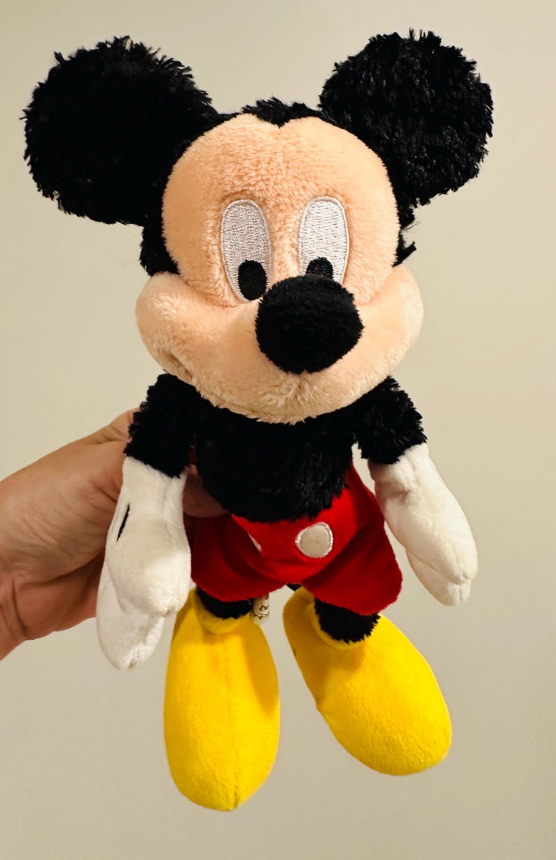 Puede incluir: Un peluche de Mickey Mouse con orejas negras, cuerpo negro, pantalones cortos rojos, zapatos amarillos y guantes blancos. El juguete tiene una cara beige y una nariz negra. El juguete es un personaje cl&aacute;sico de Disney.