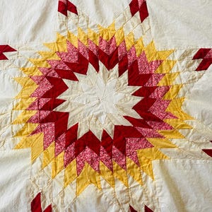May include: A handmade quilt featuring a large central star pattern composed of diamond-shaped fabric pieces in shades of red, pink floral print, and yellow, all set against a solid off-white background with visible stitched quilting lines.