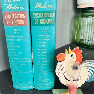 May include: Two vintage turquoise "Meta Given's Modern Encyclopedia of Cooking" books with gold lettering. Volume II covers Fo-Z, and Volume I covers A-Fi. A wooden rooster-shaped timer with a red sand timer sits in front of the books.
