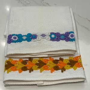 May include: Two white hand towels with decorative embroidered borders. One towel features a blue, purple, and white floral design. The other towel has a brown, orange, and yellow leaf pattern.