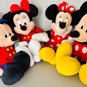 May include: Four plush Disney character dolls: Mickey Mouse and Minnie Mouse. Mickey wears a red jacket, black pants, and white gloves. Minnie wears a red dress with white polka dots and a red bow. The dolls are on a white surface.
