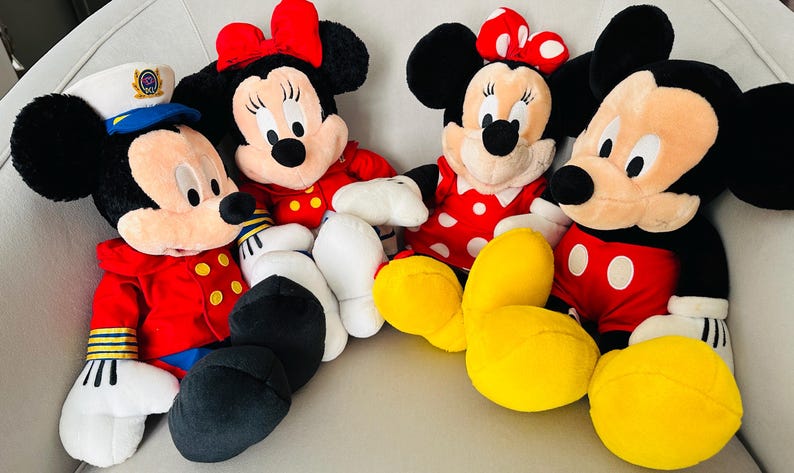 May include: Four plush Disney character dolls: Mickey Mouse and Minnie Mouse. Mickey wears a red jacket, black pants, and white gloves. Minnie wears a red dress with white polka dots and a red bow. The dolls are on a white surface.