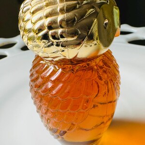 K&ouml;nnte beinhalten: Dekorative parf&uuml;mflasche in Eulenform. Der obere Teil ist goldfarben, der K&ouml;rper bernsteinfarben. Die Flasche hat eine strukturierte Oberfl&auml;che, die Federn &auml;hnelt. Augen und Schnabel der Eule sind sichtbar. Die Flasche steht auf einer wei&szlig;en Oberfl&auml;che.